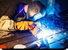 Worker wearing safety gear and welding helmet performing metal welding on a workbench, with bright sparks and blue light from the welding process.