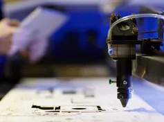 Close-up of a laser cutting machine precisely etching a design on a flat sheet, with a technician holding a tablet in the background.